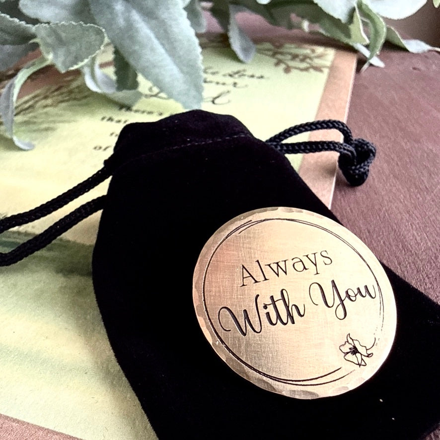 Round brass tag with 'Always With You' engraving on a black drawstring bag.