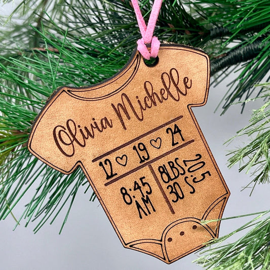 Wooden ornament shaped like a baby onesie with engraved text, hanging on a Christmas tree.