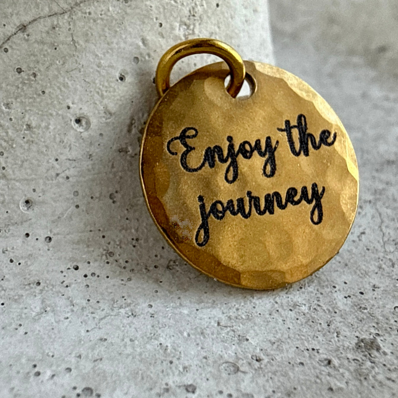 Gold pendant with 'Enjoy the journey' engraving on a concrete surface