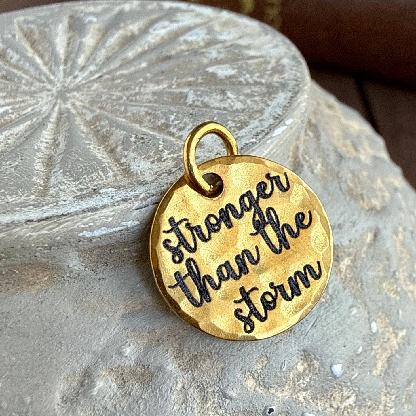 Gold pendant with 'stronger than the storm' engraving on a stone surface