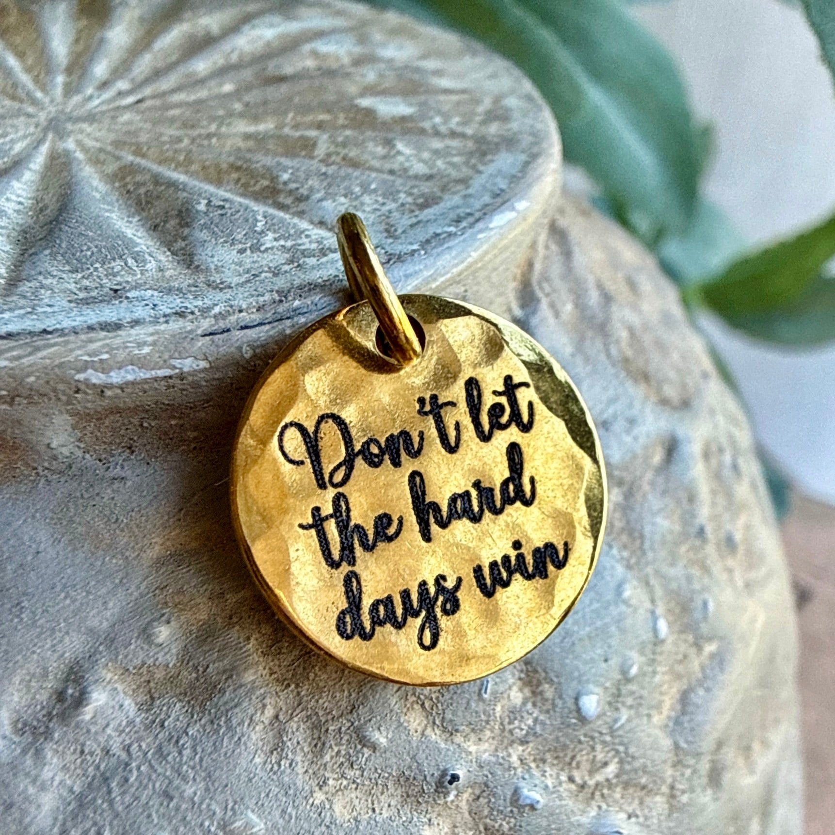Gold pendant with 'Don't let the hard days win' engraving on a textured stone surface.