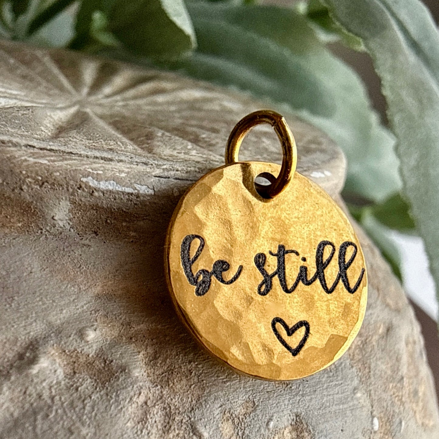 Gold pendant with 'be still' and heart on a stone surface