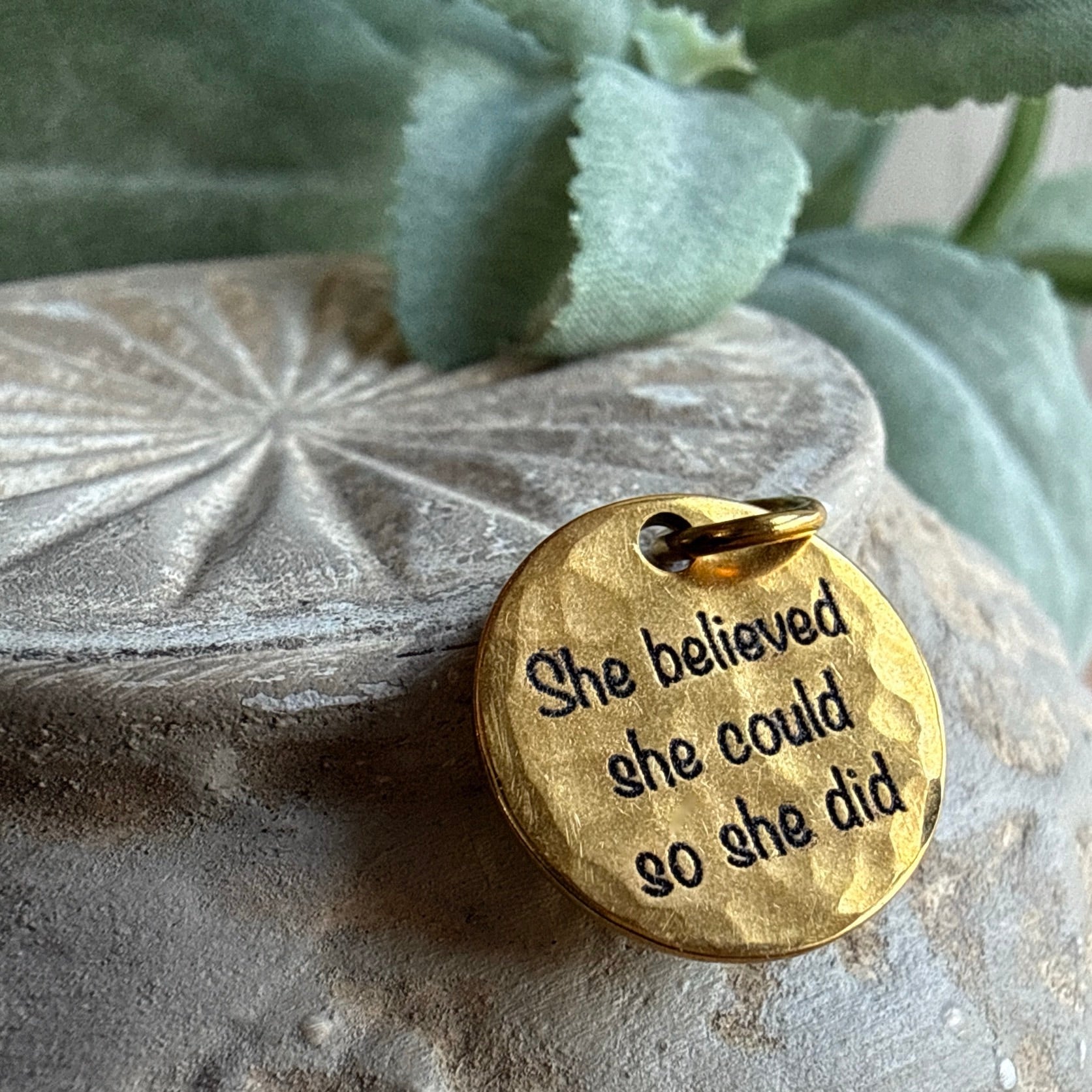 Gold pendant with 'She believed she could so she did' text on a stone surface 