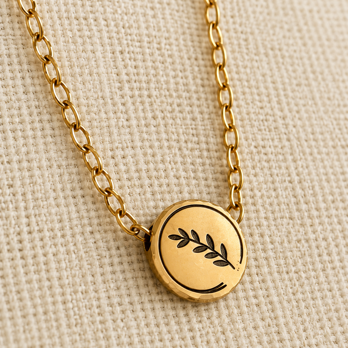 A gold chain necklace with a round gold pendant featuring a black engraved leaf motif, displayed on a textured beige fabric background.